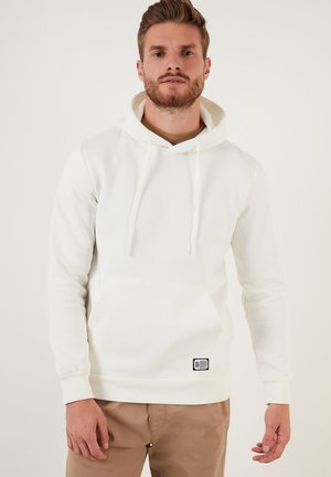 White hoodie featuring a kangaroo pocket, drawstring hood, and ribbed cuffs and hem. Small black logo tag at the lower left. Soft material.