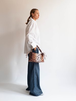 Geantă de mână cu imprimeu de leopard, cu o textură moale, curele lungi și o formă rotundă. Modelul poartă o cămașă albă și pantaloni largi din denim.