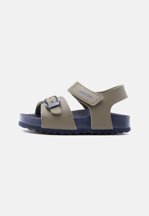 Sandaletto open-toe per bambino con due cinturini, una fibbia regolabile e una chiusura in Velcro, tomaia beige e suola blu scuro.