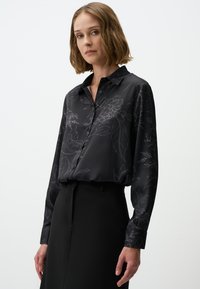 Black long-sleeve blouse featuring a floral pattern in light gray, button-down front, and a classic collar. Paired with a black skirt.