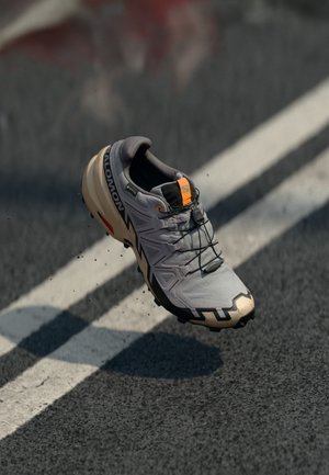 Gray athletic shoe with black accents, textured upper, and orange detail on the tongue. Features a rugged sole for traction and stability.