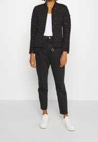 Kobieta w czarnej, cekinowej, pikowanej kurtce, białym t-shircie, czarnych dżinsach z wysokim stanem i białych trampkach na platformie stoi na tle białym.