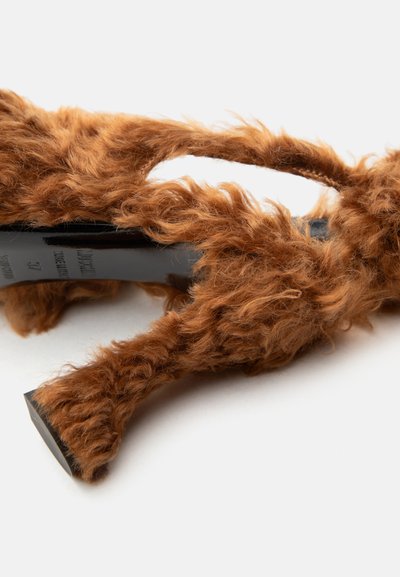 Καφέ χνουδωτό πέδιλο με ψηλό τακούνι, με χοντρές λουρίδες και ανοιχτή μύτη, σε λευκό φόντο, τοποθετημένο υπό γωνία για να φαίνονται οι λεπτομέρειες του τακουνιού και της πλευράς.