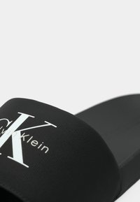Black slide sandal featuring textured upper with white branding, smooth matte footbed, and rounded edges for comfort.
