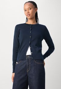 Marineblauwe gebreide cardigan met knopen aan de voorkant, geribde mouwen en onderrand, gedragen over een wit topje, gecombineerd met donkerblauwe spijkerbroek.