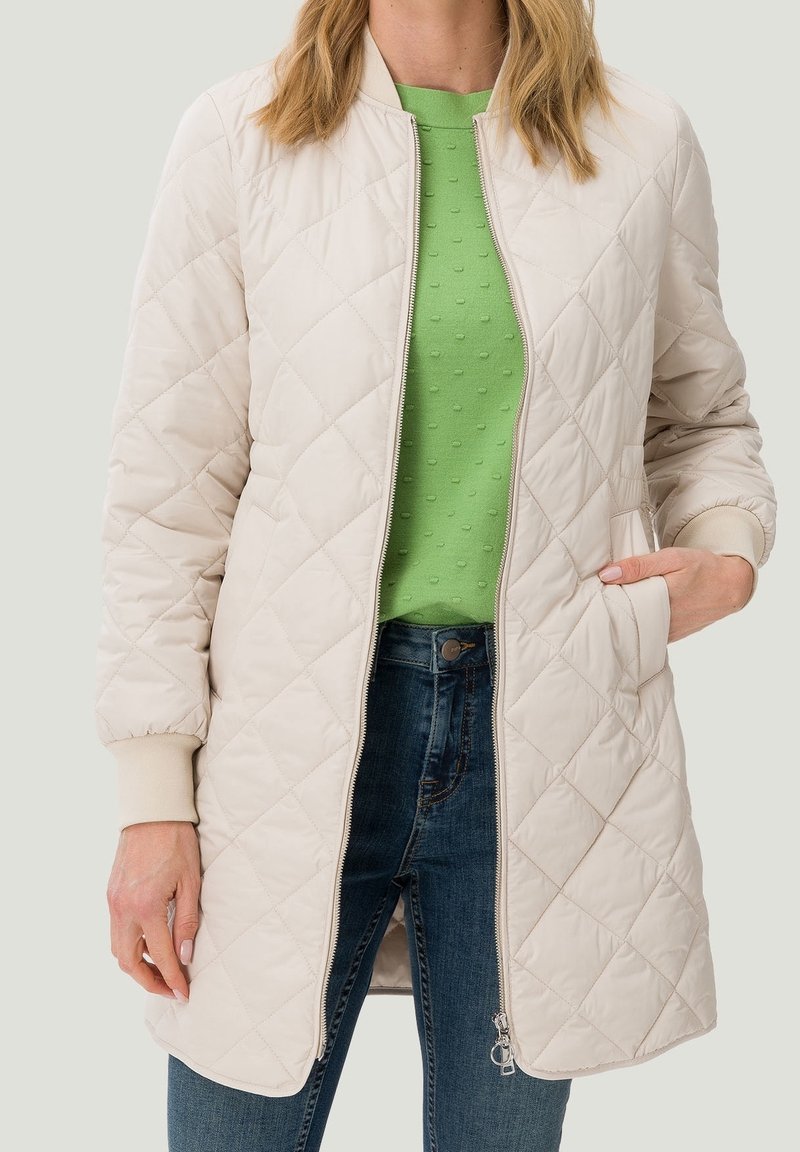 Giacca trapuntata beige in stile bomber, con zip frontale, polsini a costine e tasche laterali, abbinata a una maglia verde testurizzata.