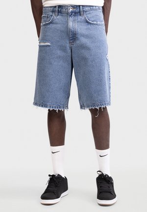 Person som har på seg lyseblå slitte jeansshorts, hvite Nike-kortesokker og svarte joggesko mot en ensfarget bakgrunn.