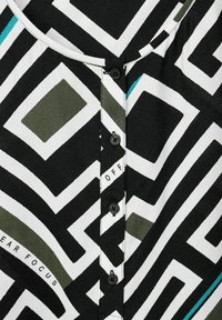 Camicia a fantasia bianco e nero con disegni geometrici, caratterizzata da un collo rotondo e bottoni neri lungo la chiusura frontale.