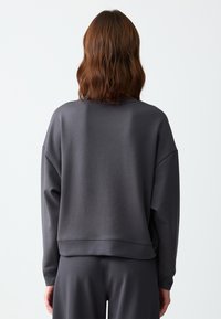 Felpa grigia sovradimensionata con spalle scese, texture morbida e orlo a costine; abbinata a pantaloni coordinati, vista da dietro.