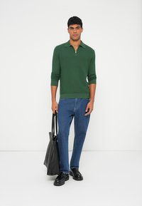 Zelený pletený pulóver s golierom na zips, v kombinácii s modrými džínsami. Modelka drží čiernu tašku a má na sebe čierne topánky. Minimalistické pozadie.