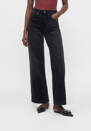Woman wearing high-waist wide-leg black jeans, one hand in pocket, paired with black pointed flats featuring metallic buckle details.