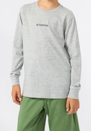 Person iført en lysegrå langærmet skjorte med "Champion" logo og løse grønne bukser, stående med arme afslappet ned langs siderne.