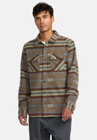 Camicia di flanella multicolore marrone con strisce orizzontali, caratterizzata da motivi grigi, blu e rossi, e due tasche frontali con pattine.