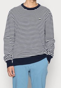 Man wearing a navy and white striped long-sleeve sweater with ribbed cuffs and hem, and light blue pants with one hand in pocket.