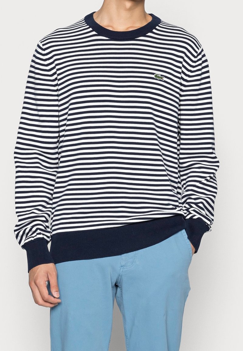 Man wearing a navy and white striped long-sleeve sweater with ribbed cuffs and hem, and light blue pants with one hand in pocket.