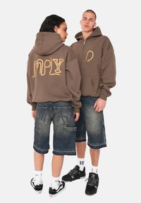 Braun kapuzen Sweatshirt mit Rope-Style-Text auf dem Rücken, kombiniert mit dunklen Jeansshorts mit gestickten Akzenten. Gezeigt werden Sneakers und Socken.