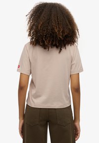 Kortærmet, lys beige T-shirt med rund hals og diskrete sømme, med en lille rød logo på venstre ærme. Glat stoftekstur.