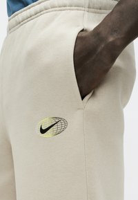 Άτομο με σκούρο δέρμα που φοράει μπεζ φόρμα με μαύρο και κίτρινο λογότυπο Nike, με το χέρι να τοποθετείται χαλαρά στην δεξιά μπροστινή τσέπη.