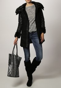 Abrigo largo negro con ribete de piel sintética, mangas texturizadas, suéter gris, jeans azules y botas negras hasta la rodilla. Sostiene un bolso tote metálico negro.