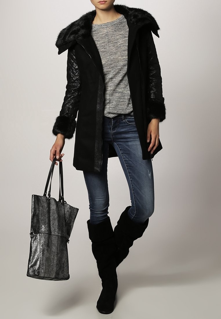 Abrigo largo negro con ribete de piel sintética, mangas texturizadas, suéter gris, jeans azules y botas negras hasta la rodilla. Sostiene un bolso tote metálico negro.