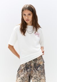 T-shirt en coton blanc à manches courtes et col rond, avec un logo rose brodé. Porté avec un pantalon large à motifs.