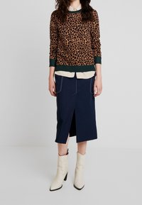 Pull en maille imprimé léopard avec des accents verts, superposé sur une chemise crème. Jupe midi marine avec fente sur le côté et coutures blanches, associée à des bottines crème.