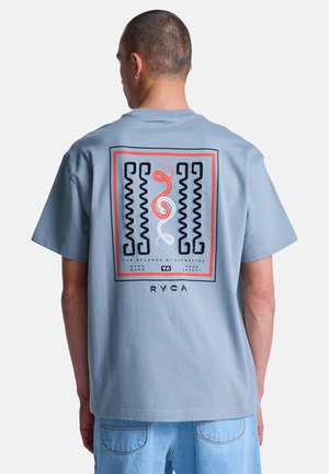 Man draagt een lichtblauw T-shirt met een zwart-rood abstract slangenontwerp en de tekst "Het evenwicht van tegenstellingen" op de achterkant.