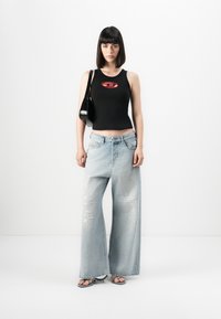 Top sans manches noir avec logo rouge, associé à un jean large déchiré bleu clair et des sandales noires. Le modèle tient un petit sac noir.