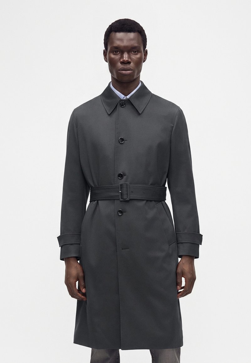 Homme portant un trench-coat gris foncé ceinturé avec un col et une fermeture à boutons, debout devant un fond clair uni.