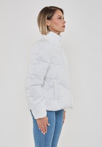 Giacca a vento bianca con collo alto, caratterizzata da un design trapuntato, tasche laterali e chiusura frontale con zip. Texture leggermente lucida.