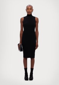 Rochie midi neagră, cu design cu nervuri, gât înalt, siluetă ajustată și fără mâneci. Purtată cu pantofi negri și o geantă mică neagră.