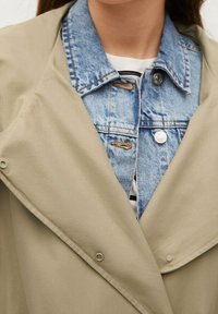 Trench-coat beige avec un large col superposé sur une veste en denim clair. Il présente des boutons en bronze et des détails de couture visibles.