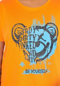 Orange kortærmet T-shirt med grafisk tekst i sort og blå samt sølv accents. Teksten lyder "NERDY DIRTY INKED AND WILD."