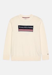 Maglione color crema realizzato in tessuto lavorato a maglia, con un rettangolo nero su cui è scritto "Tommy HILFIGER" in testo bianco e con strisce rosse e blu.