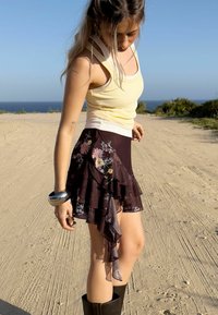 Jupe noire à volants avec imprimé floral dans des teintes de rose et de jaune ; design superposé ; portée avec un débardeur jaune clair et des bottes noires.