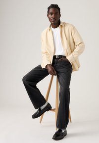 Φωτεινό μπεζ πουκάμισο με κουμπιά πάνω από λευκό t-shirt, συνδυασμένο με σκούρο, άνετο τζιν τζιν και μαύρα μοκασίνια. Απλός ξύλινος σκαμπό.