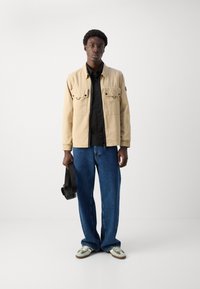 Beige lichtgewicht jack met twee voorzakken, zwart overhemd met knopen, losse blauwe denim jeans en crèmekleurige sneakers met groene accenten.