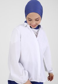 Hvid langærmet skjorte med knaplukning foran, blødt stof og elastiske ærmekanter. Båret med en blå hijab.