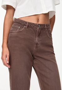 Pantalon en denim marron avec une coupe décontractée, doté d'une fermeture à bouton et à glissière, de poches avant et d'une texture subtile. Chemise raccourcie visible au-dessus.