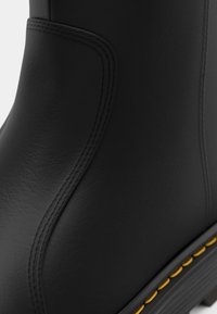 Botín de goma negro con textura suave, diseño curvado y gruesos pespuntes amarillos a lo largo de la suela. Presenta una parte superior de corte alto con costuras detalladas.