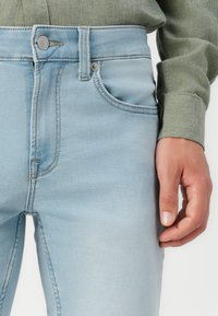 Vaqueros de mezclilla azul claro de cintura alta, con un único bolsillo frontal, botón metálico y detalles de costura sutiles. Mano visible en el lado.