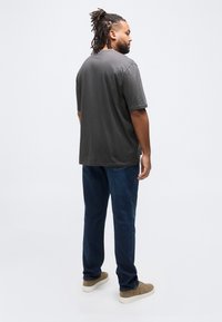 Άνδρας με dreadlocks που στέκεται, κοιτάζοντας ελαφρώς προς τα δεξιά, φοράει σκούρο γκρι T-shirt, μπλε τζιν και καφέ σουέντ αθλητικά παπούτσια σε λευκό φόντο.