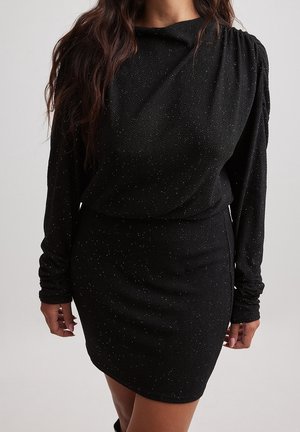 Kobieta nosi czarną mini sukienkę z długim rękawem, o fakturowanej tkaninie, delikatnie połyskującą i ze zmarszczonymi rękawami, stojąc na jednolitym jasnym tle.