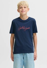 Plavi dječak s plavom majicom kratkih rukava s crvenim i bijelim tekstom "Jack & Jones", u kombinaciji s svijetloplavim trapericama, stoji ispred bijele pozadine.