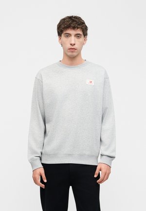 Junger Mann mit lockigem Haar, der einen hellgrauen Sweatshirt mit einem kleinen Logopatch auf der Brust trägt, steht vor einem schlichten weißen Hintergrund.