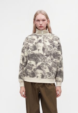 Personne portant un pull polaire à col montant avec un motif abstrait gris et blanc et un pantalon marron sur un fond clair uni.