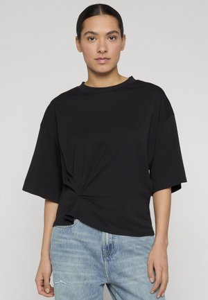 Kobieta o ciemnych włosach, związanych w niski koczek, ubrana w luźną czarną bluzkę z krótkim rękawem z detalem skręconego węzła oraz jasnoniebieskie dżinsy.