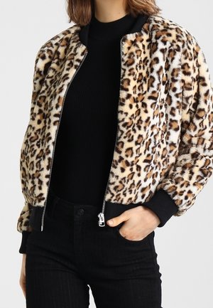 Personne portant une veste zippée imprimé léopard par-dessus un col roulé noir et un pantalon noir, avec une main dans la poche.