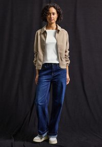 Veste bomber beige sur une chemise blanche, associée à un jean large bleu marine et des sneakers clairs. Tissu lisse, coupe décontractée et silhouette droite.