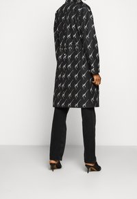 Osoba ubrana w czarny długi płaszcz z białym wzorem, czarne proste spodnie i czarne obcasy, stojąca na jednolitym tle.